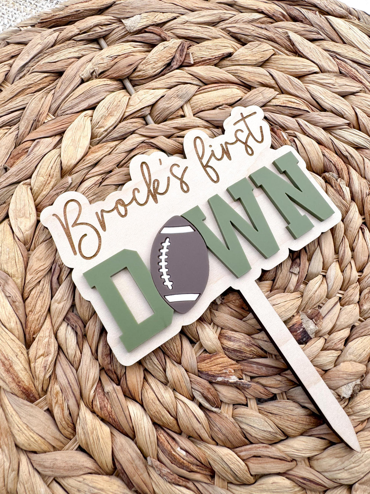 Close-up of a personalized First Down cake topper showing sage green layered acrylic DOWN lettering, a brown acrylic football accent and an engraved name on a natural maple wood backer