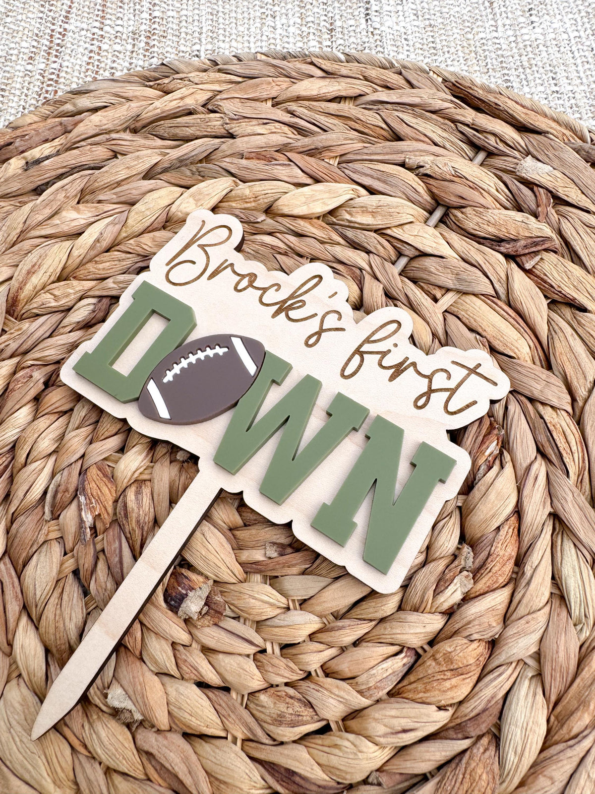 Angled view of a sage green First Down cake topper showing the dimensional layered acrylic letters, football accent and engraved script name on the wood backer