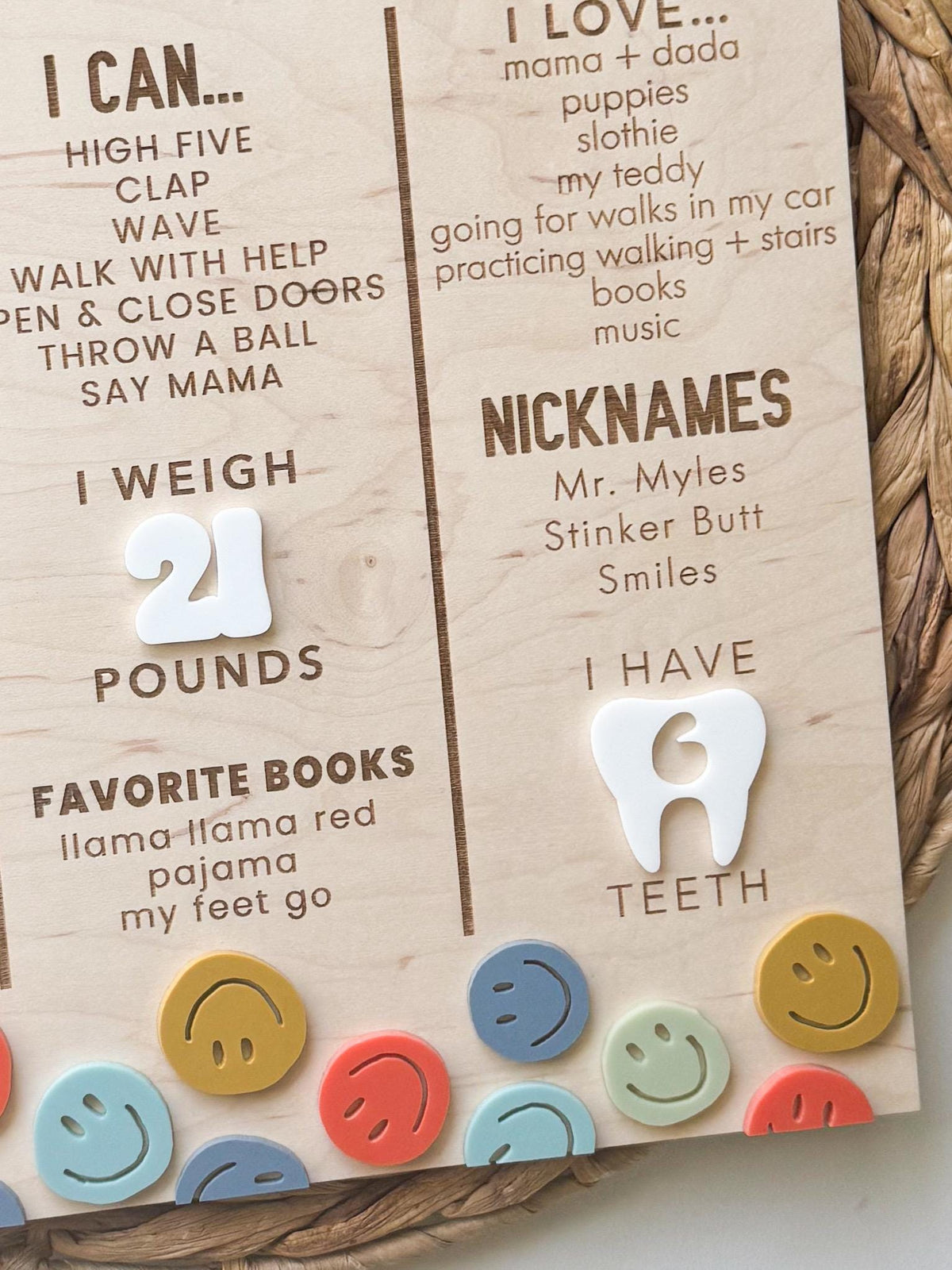 Close-up of engraved milestones on a first birthday board, including weight, nicknames, favorite books and a white acrylic tooth showing number of teeth, above layered smiley faces