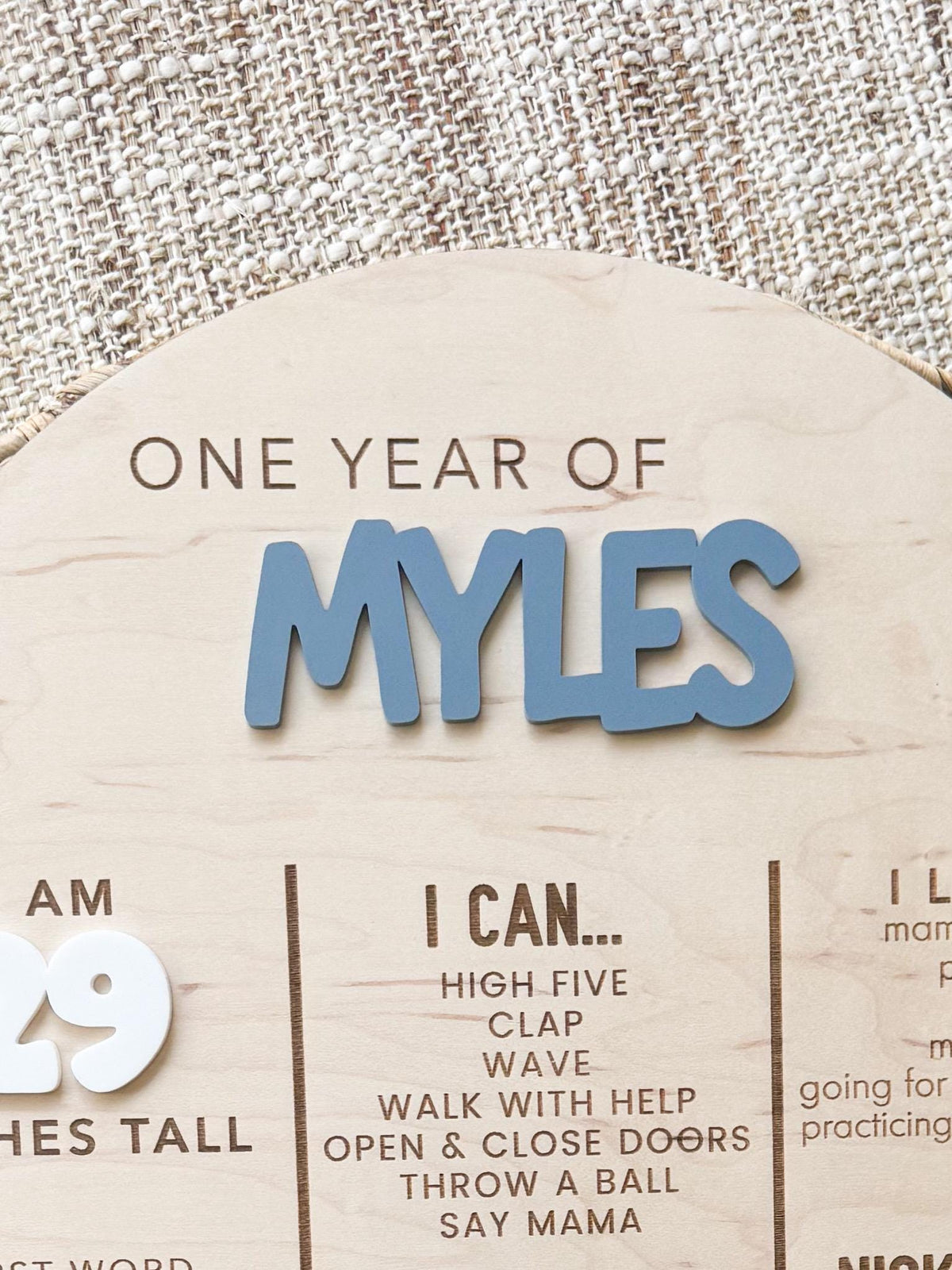Close-up of the One Year Of heading with a dimensional blue acrylic name and engraved I Can milestone list on a maple wood birthday board