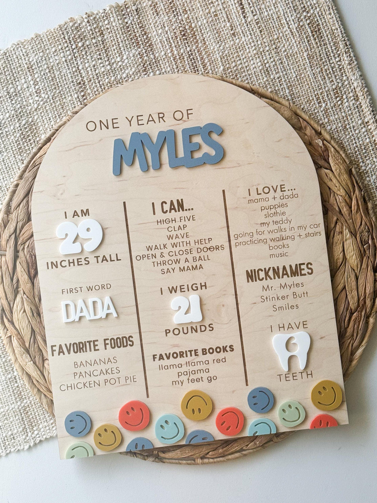 Smiley face first birthday stats board displayed on a woven placemat, showing height, weight, first word, favorite foods and the things baby loves