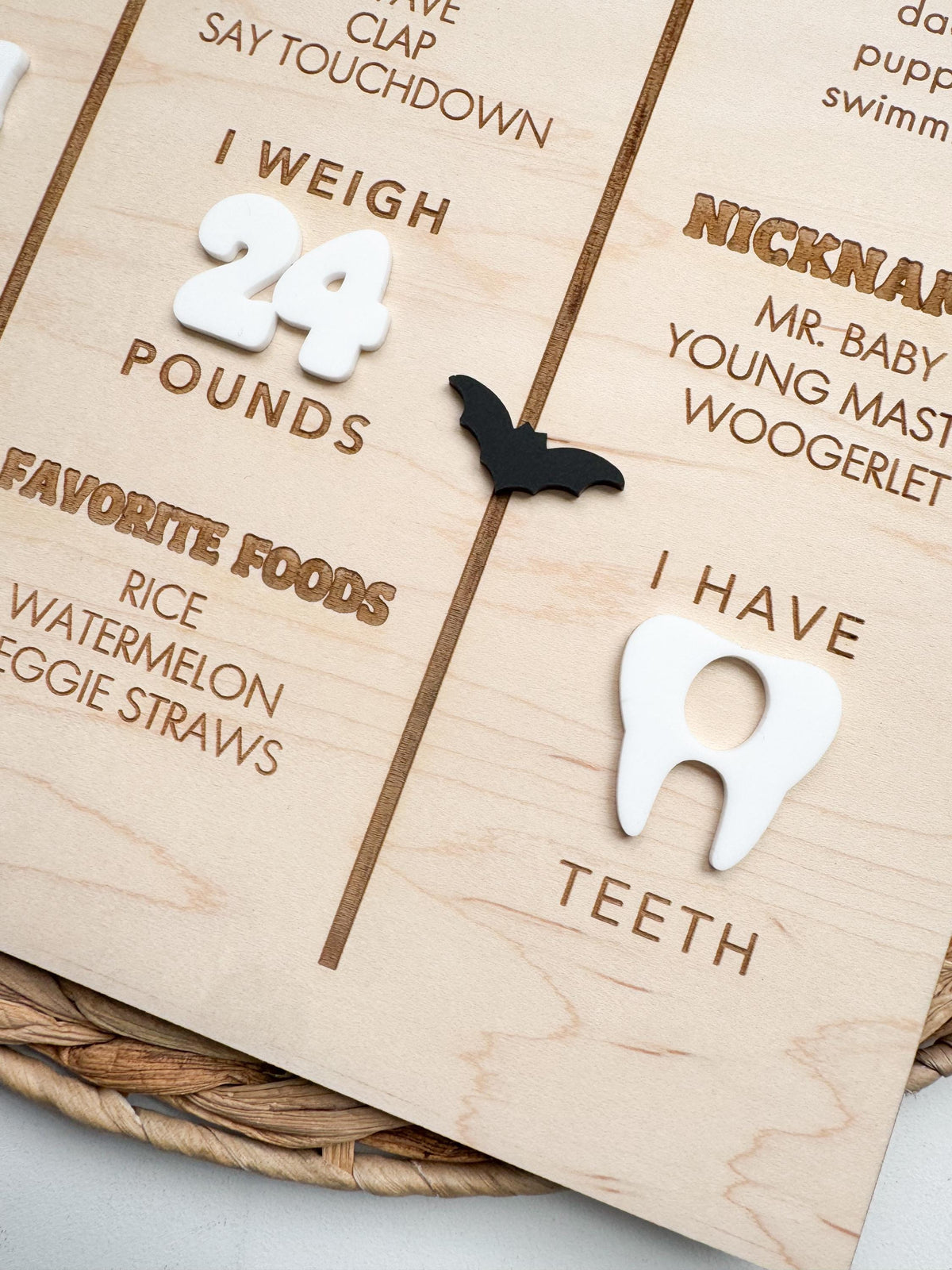 Close-up of white acrylic tooth and black acrylic bat on a baby first birthday milestone board, with laser engraved I Have Teeth, I Weigh, favorite foods and nicknames text on natural maple wood.