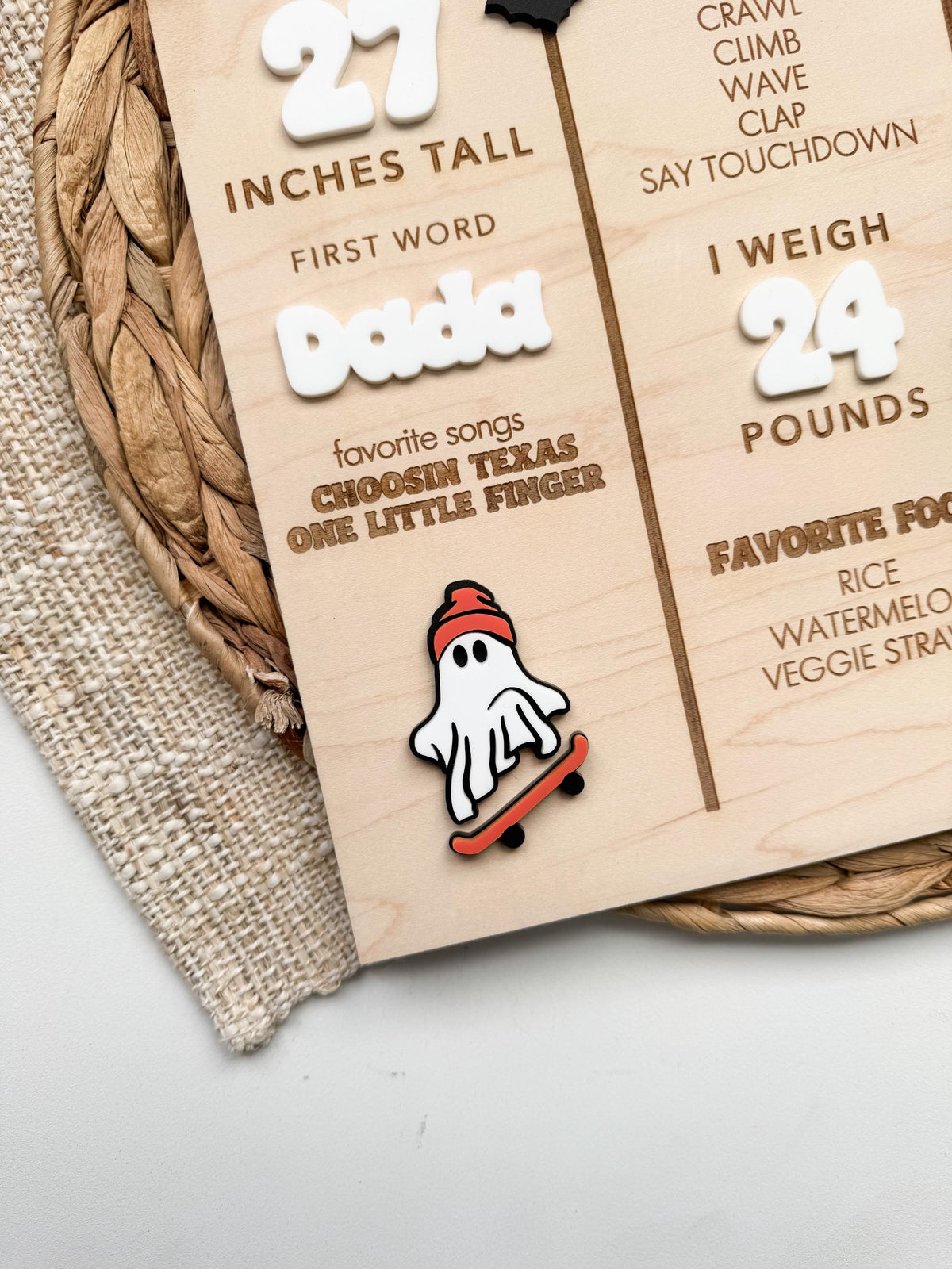 Detail of layered acrylic skateboarding ghost wearing an orange beanie on a Halloween baby milestone sign, with white acrylic first word, height and weight numbers and engraved favorite songs and foods on maple wood.