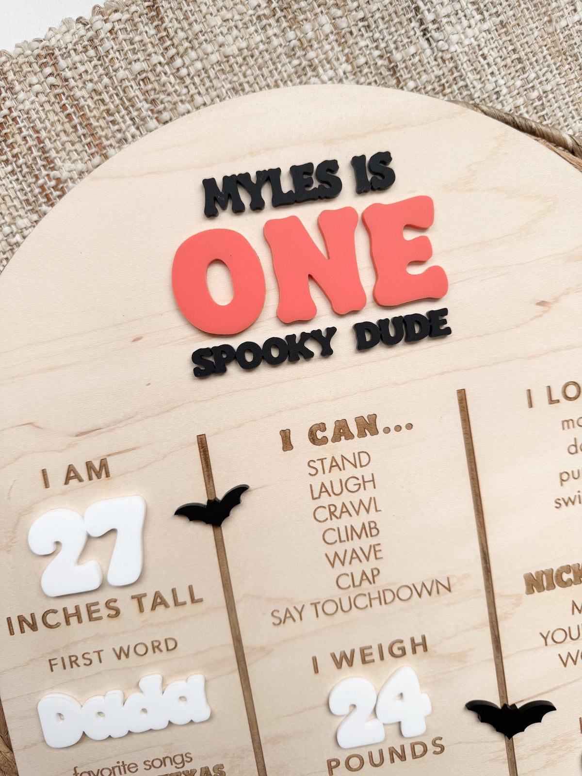 Close-up of personalized Halloween first birthday sign with layered acrylic name, coral orange ONE and Spooky Dude lettering, black acrylic bats, white acrylic height and weight numbers and engraved milestone text on maple wood.