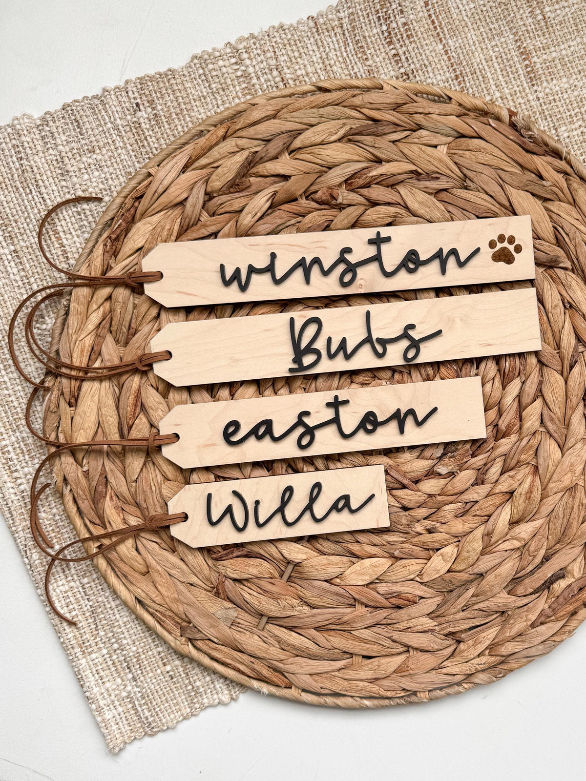 Four personalized maple wood stocking name tags with brown ties on a woven placemat, including a pet tag with paw prin