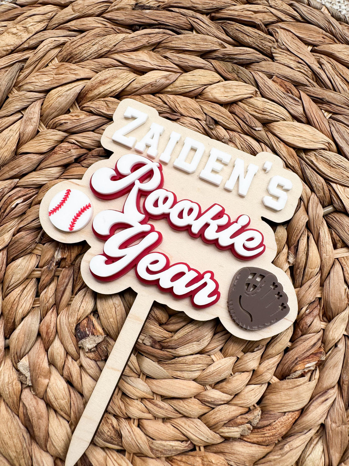 Angled view of a personalized baseball first birthday cake topper with a custom name, retro Rookie Year lettering, a baseball and a brown baseball glove, attached to a wood cake stake.