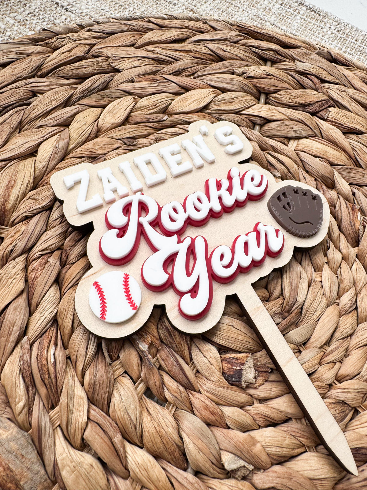 Side angle of a dimensional Rookie Year baseball cake topper showing raised acrylic layers, custom name, baseball and glove accents for a baseball themed 1st birthday party.