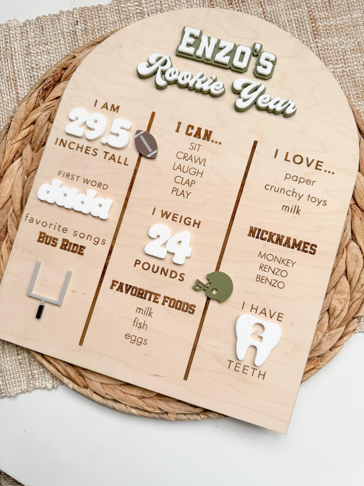 Angled view of a handmade Rookie Year first birthday milestone board with an arched maple wood base, layered white and green acrylic lettering, engraved baby stats and acrylic football, helmet, goal post and tooth details, a football birthday sign that doubles as a first year keepsake