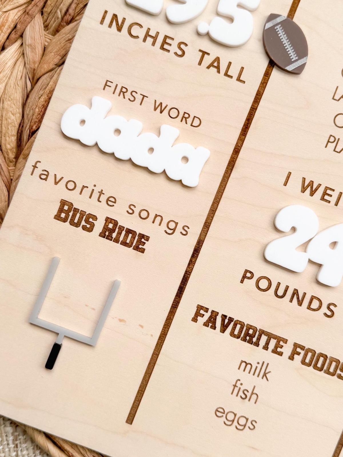 Close-up of the engraved and layered details on a football first birthday milestone board, showing a white acrylic first word, engraved favorite song, a gray and black acrylic goal post, white acrylic weight numbers and an engraved favorite foods list on natural maple wood