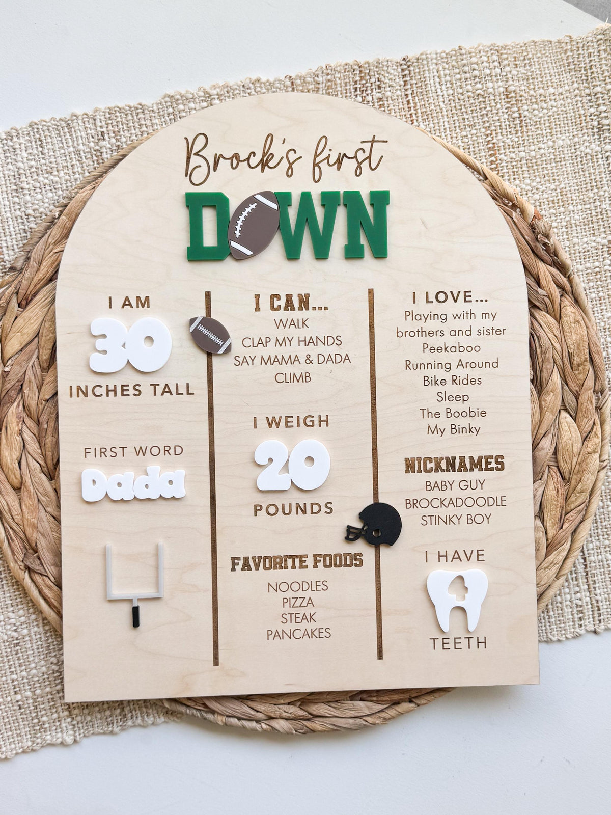 Overhead view of a First Year Down football milestone board for a first birthday party, with a personalized name, green and brown acrylic title, white acrylic stats and engraved favorites on an arched maple wood sign resting on a woven placemat