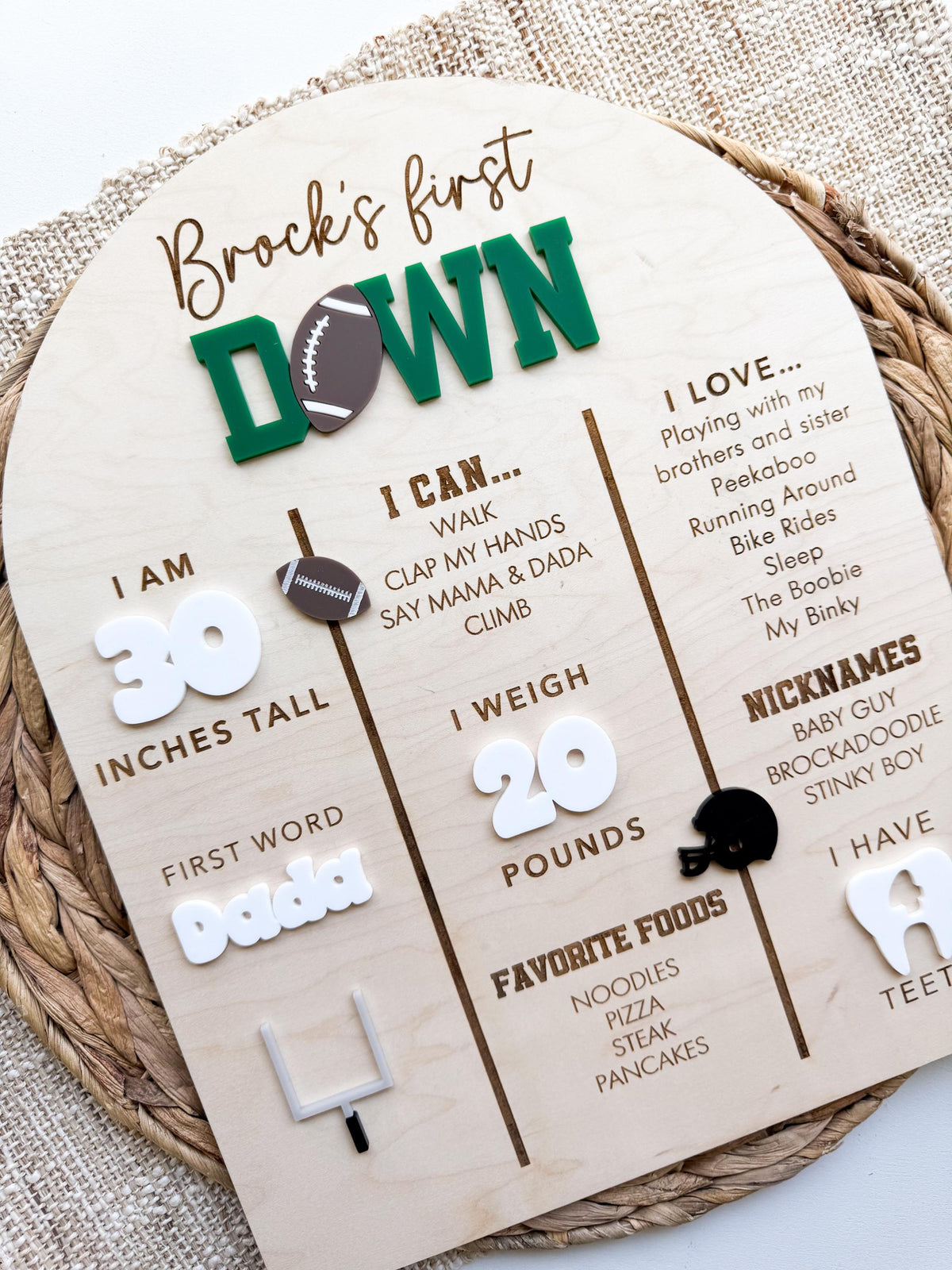 Angled view of a football first birthday stat board in maple wood with a custom name and First Down title in green and brown acrylic, white acrylic milestone numbers, engraved baby stats and a black acrylic helmet, styled on a woven placemat as first birthday party decor