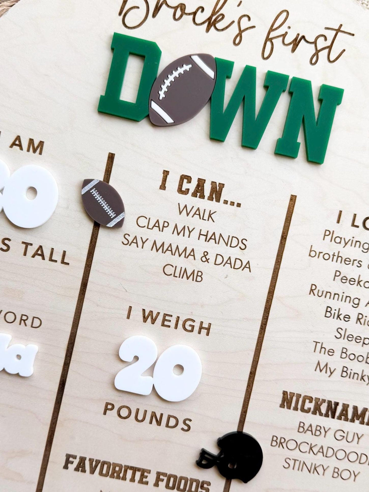 Detail of the green acrylic DOWN lettering with a brown acrylic football, a laser cut football accent, engraved I can skills, white acrylic weight numbers and a black acrylic helmet on a first birthday milestone board made from maple wood