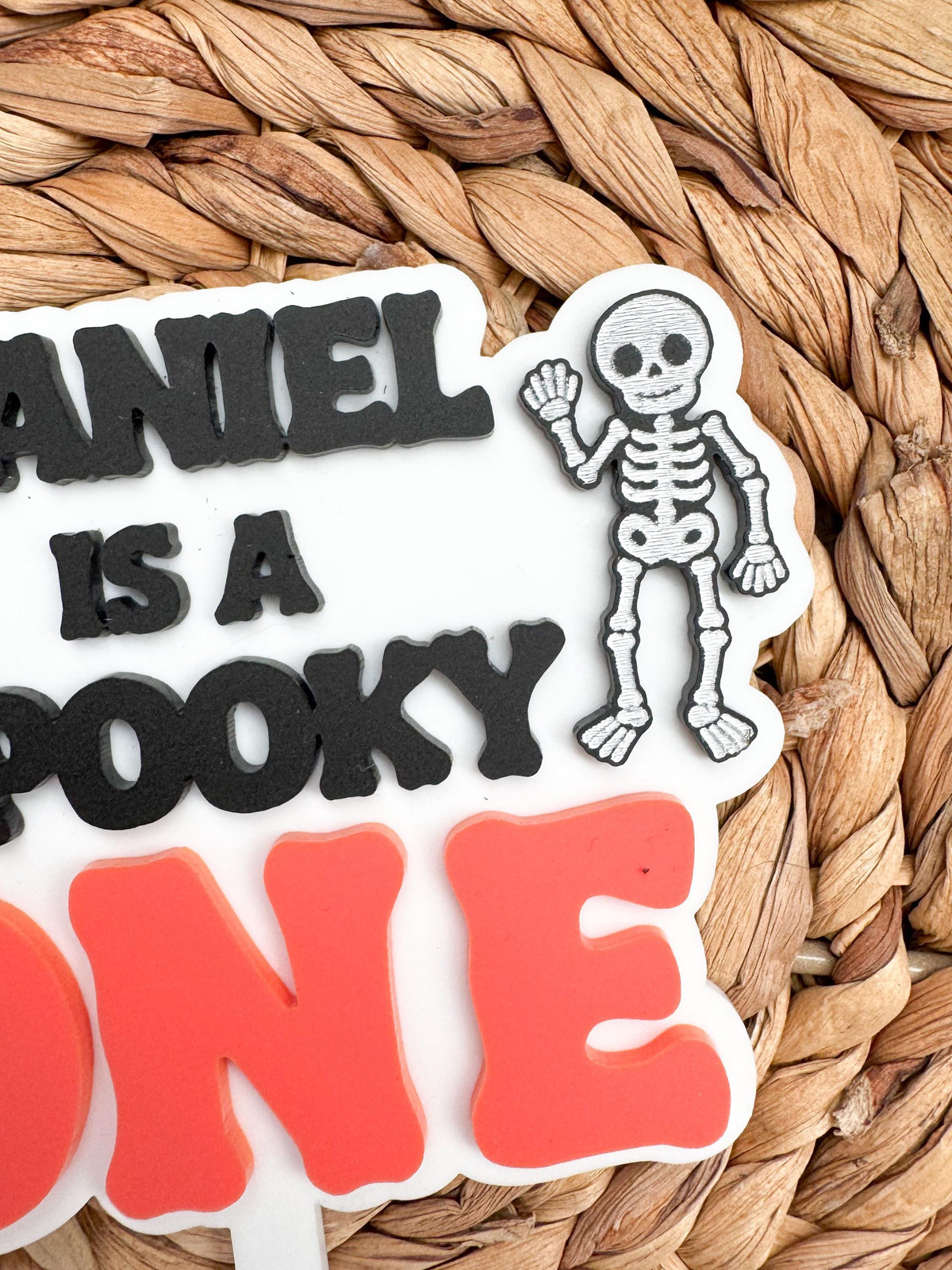 Close-up of a layered acrylic Spooky One cake topper showing raised black lettering, a waving skeleton accent and bold coral orange ONE for a Halloween first birthday.