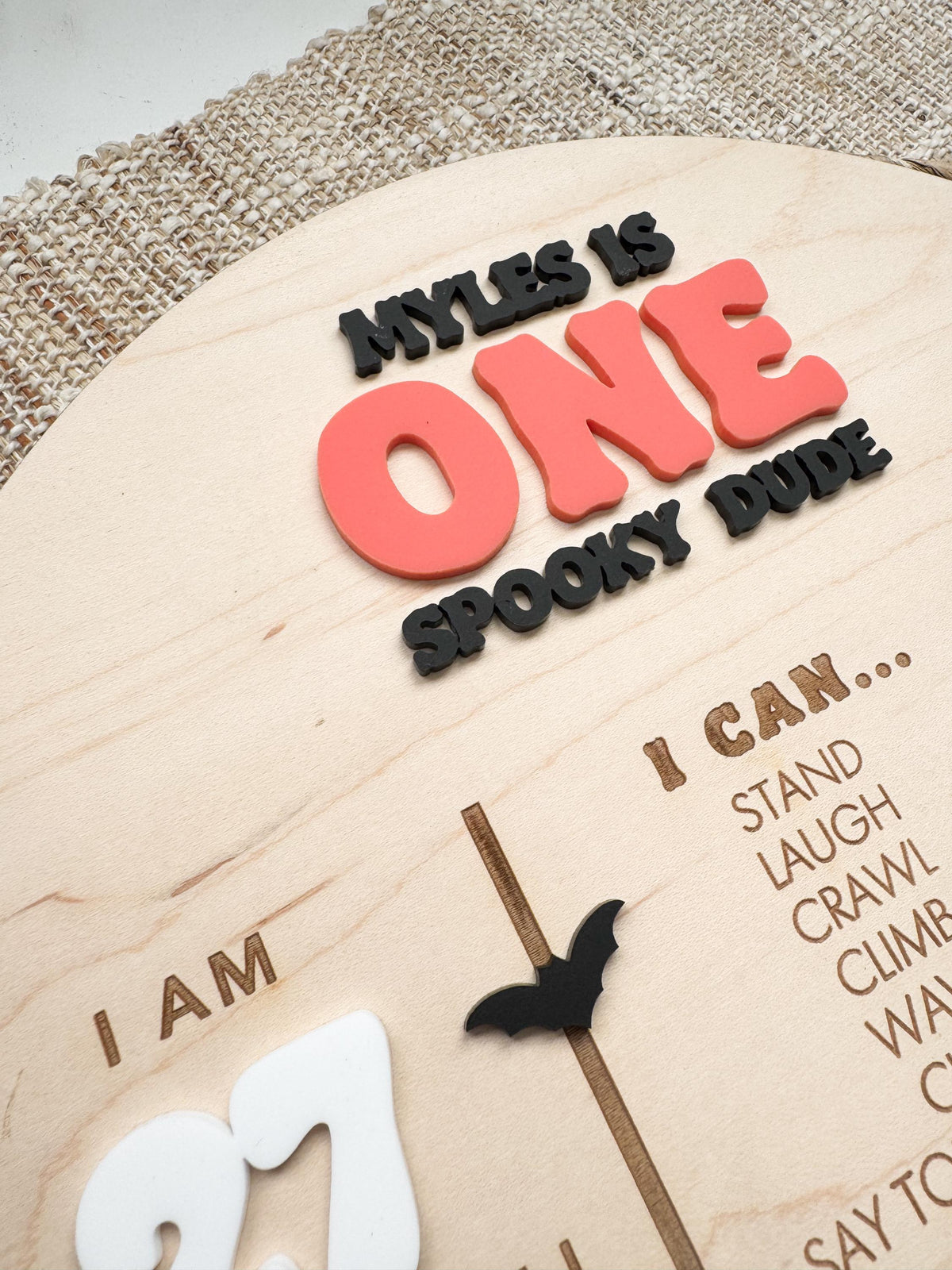 Angled detail of dimensional layered acrylic lettering on One Spooky Dude birthday sign, showing raised coral orange ONE, black acrylic text and bat, and laser engraved I Can milestone list on natural maple wood.