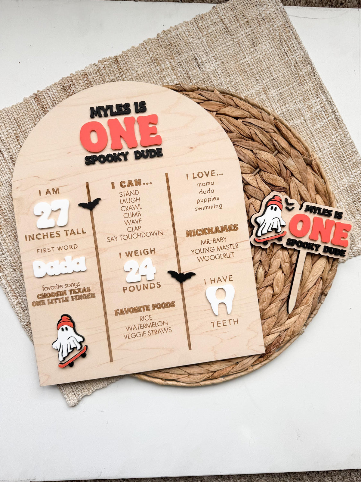 Ghost milestone sign and matching One Spooky Dude cake topper on a woven placemat, coordinating Halloween first birthday party decor with layered acrylic ghost, ONE lettering and engraved maple wood.
