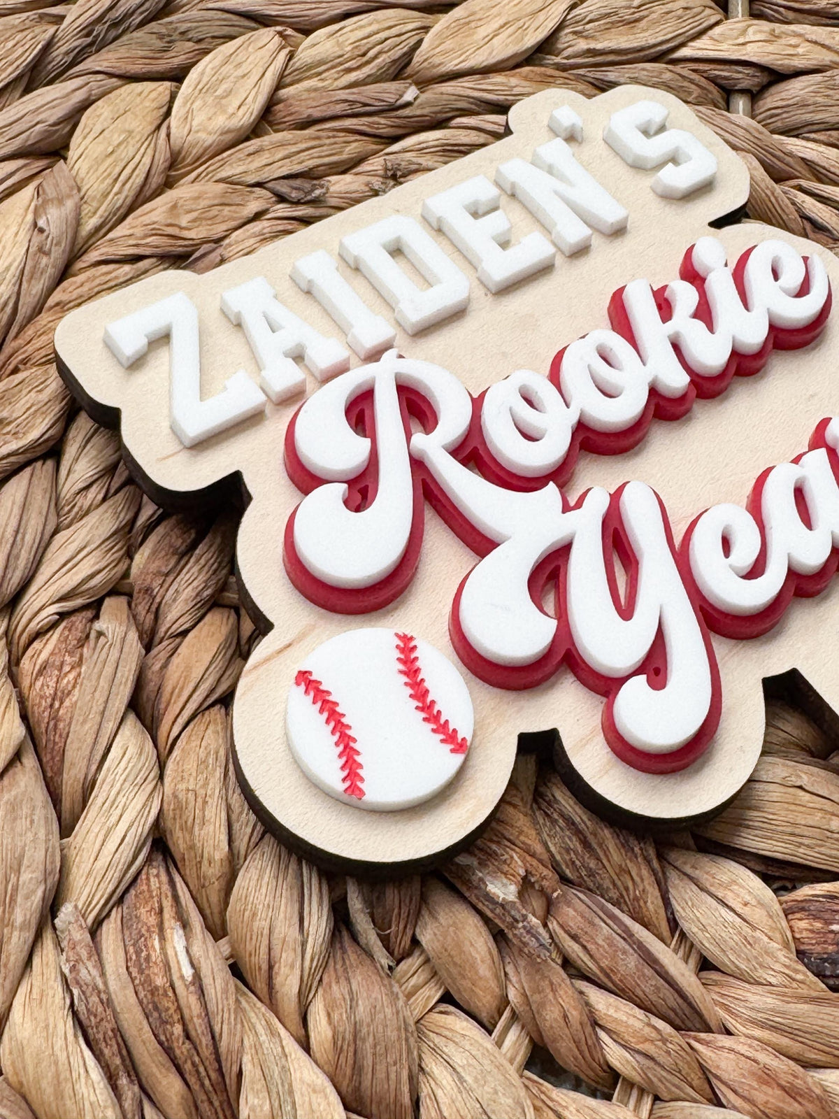 Close-up of dimensional acrylic Rookie Year script lettering in white with a red outline and a baseball accent on a personalized wood baseball first birthday cake topper.