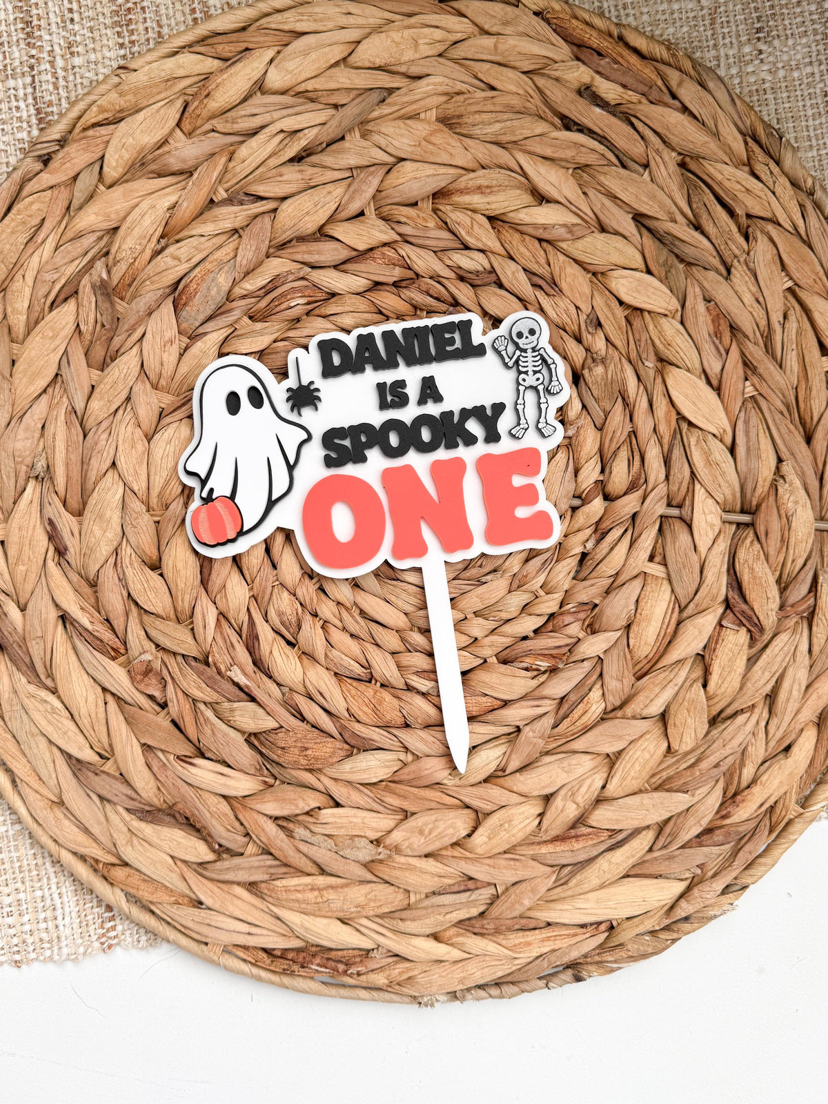 Personalized Spooky One first birthday cake topper featuring a white ghost holding an orange pumpkin, black spider, skeleton and coral orange ONE lettering layered on a white acrylic backer.