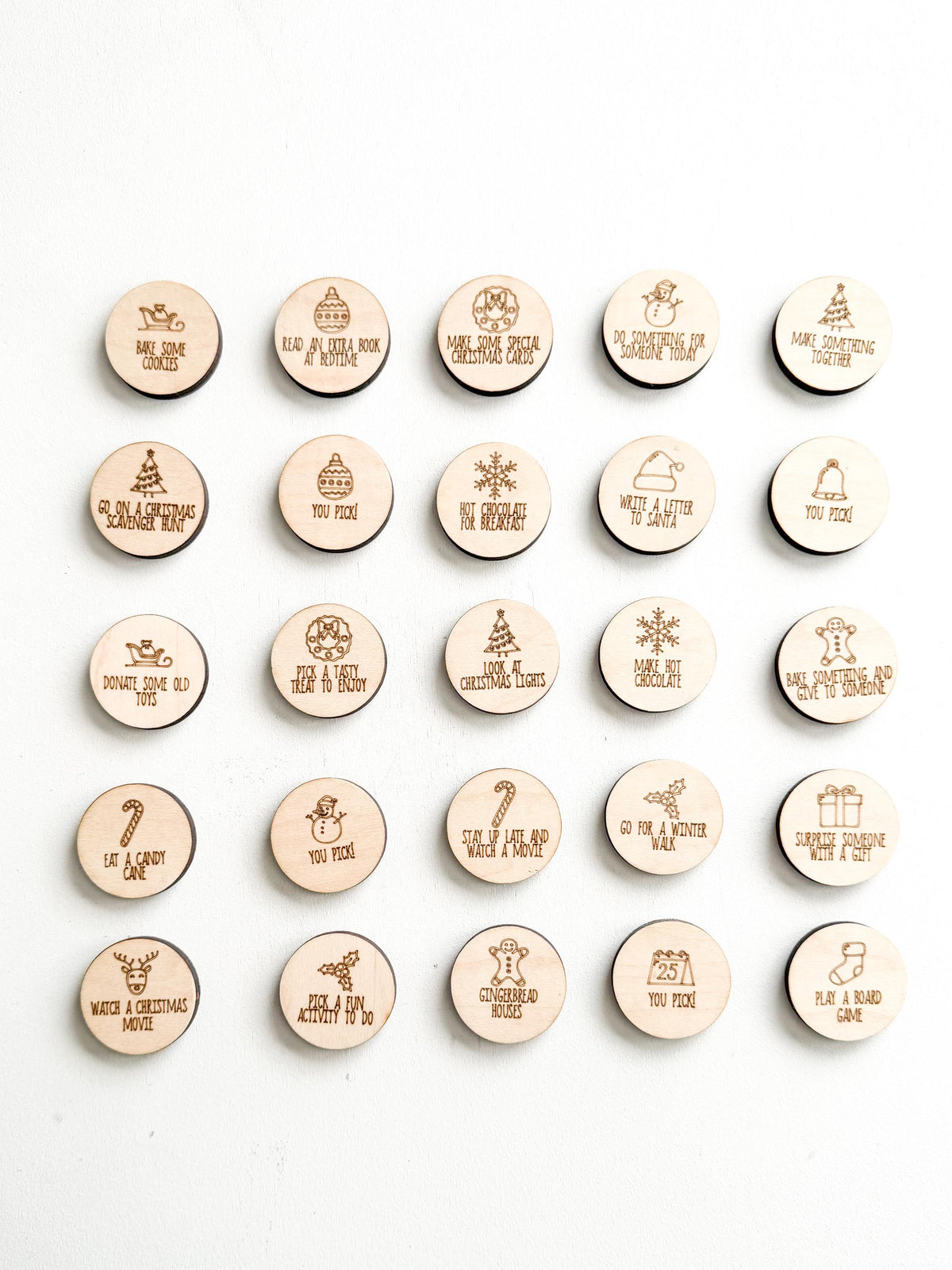 All 25 wooden Christmas advent calendar tokens arranged in five rows, showing every engraved activity in the standard set, such as Bake Some Cookies, Make Hot Chocolate, Go for a Winter Walk and Play a Board Game.